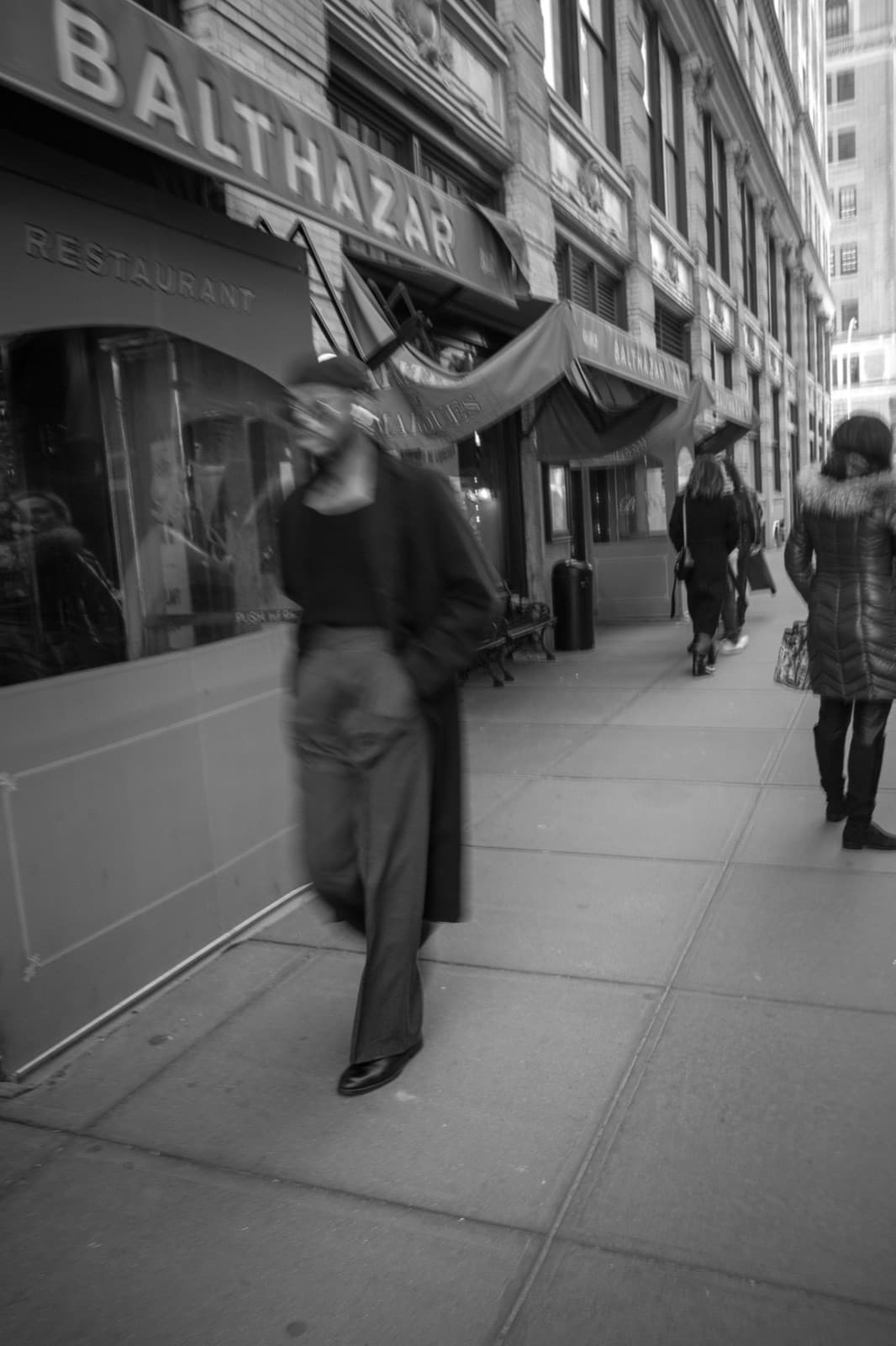 A suited pedestrian passes Balthazar on a busy New York sidewalk in black and white.