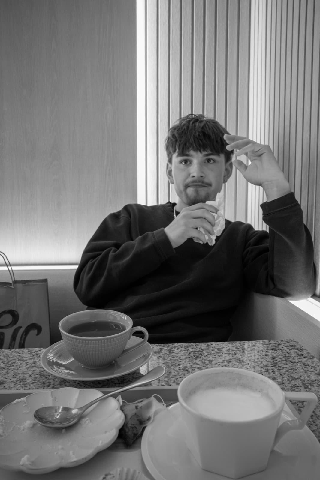 A young man gestures across a cafe table between two cups in black and white.