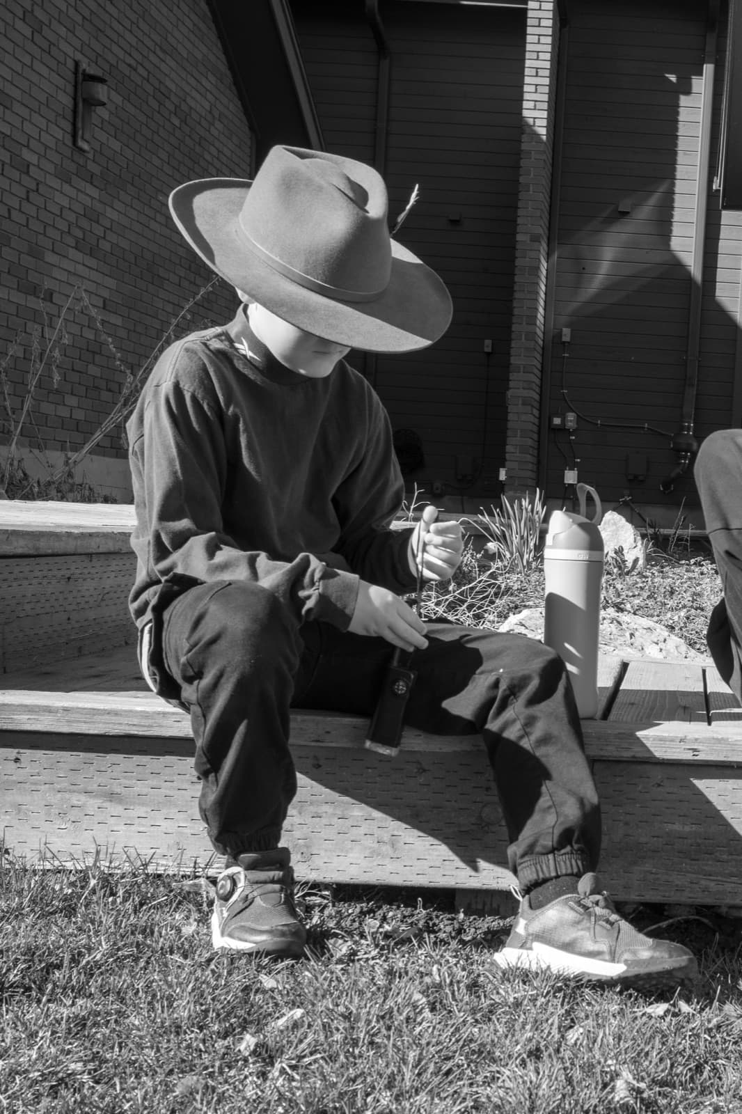A man in a broad hat sits outdoors with a thermos at his side.