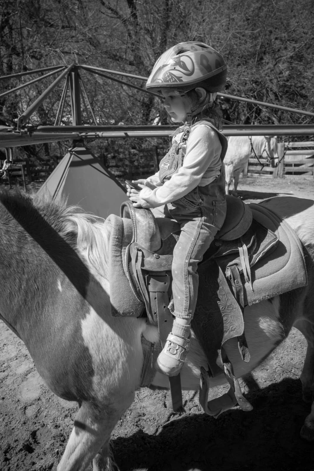A helmeted child rides a pony past a wooden corral in black and white.