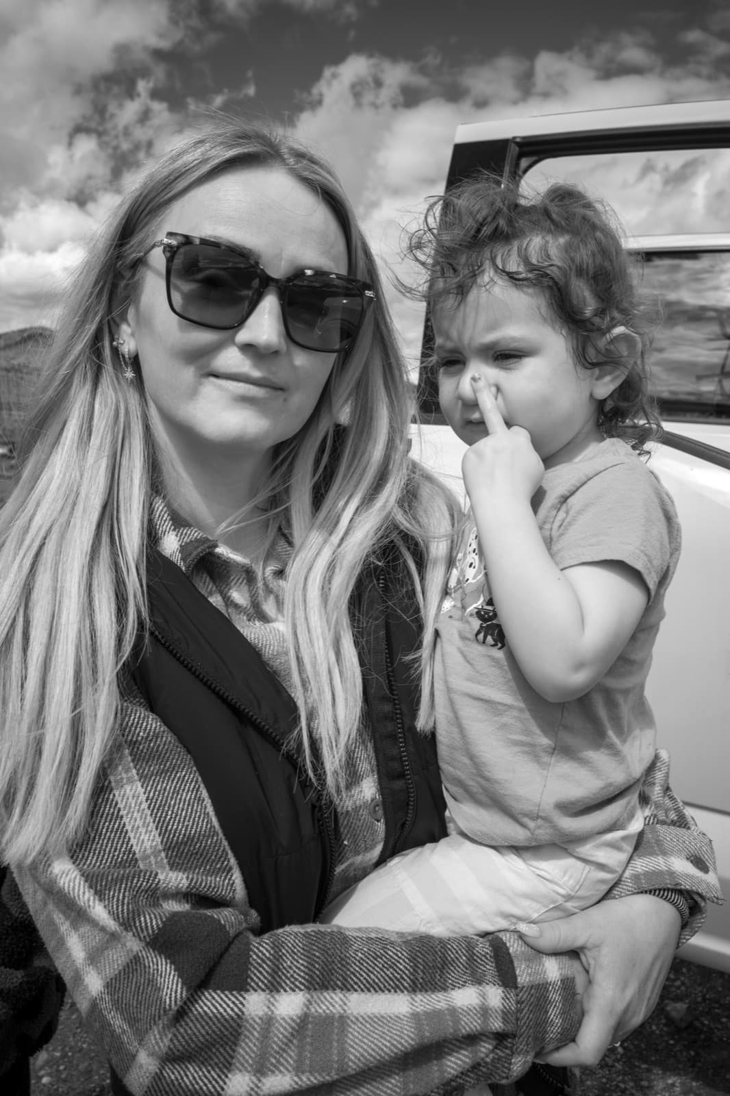 A woman in sunglasses holds a young child outdoors beneath a bright sky.