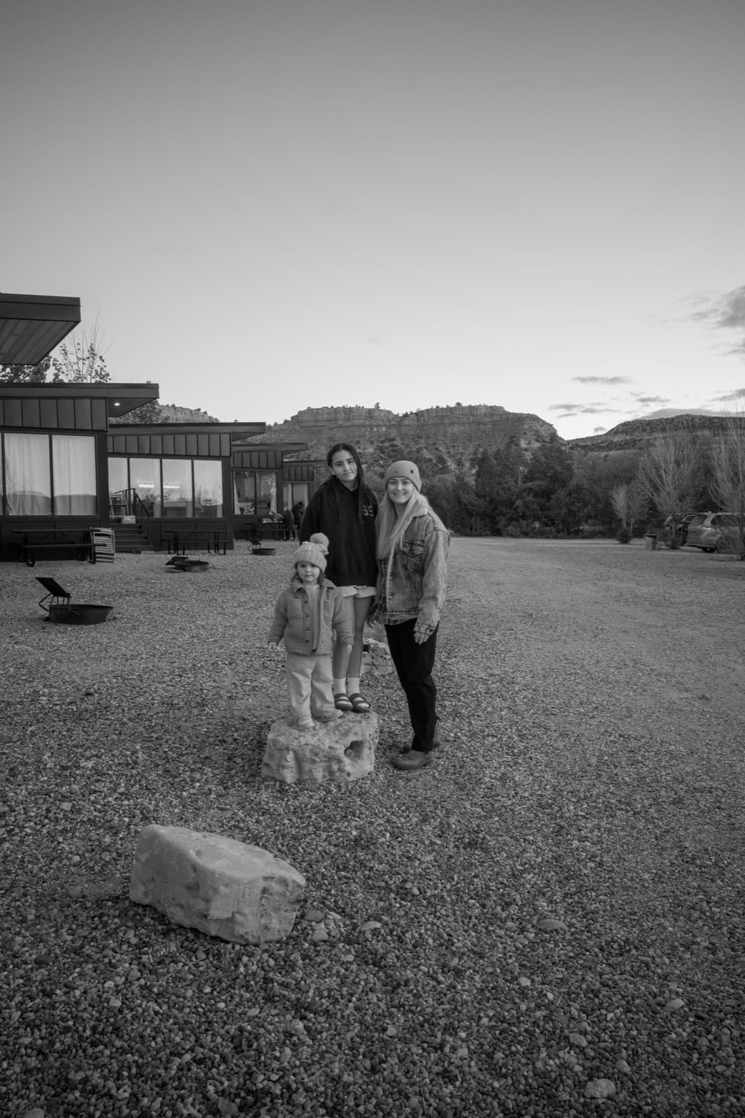 Two adults, a child, and a dog stand together in a wide gravel ranch yard.