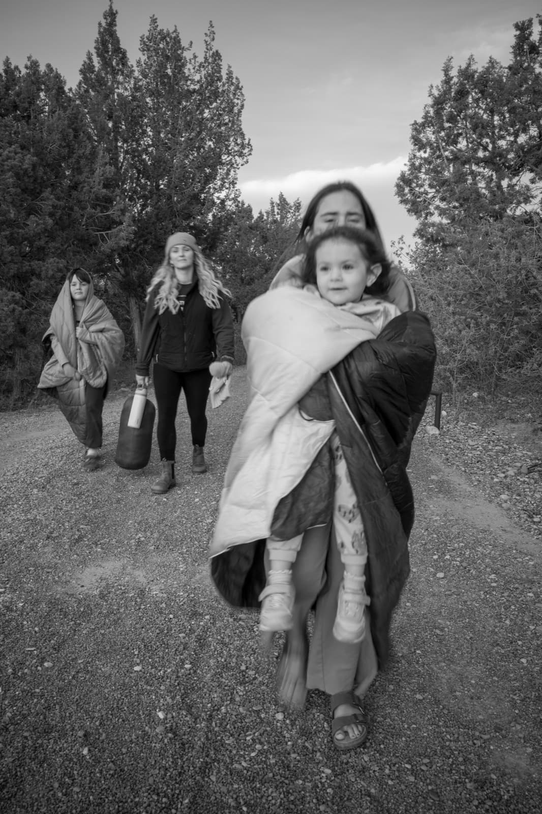 A small group walks a dirt trail while one adult carries a bundled child.