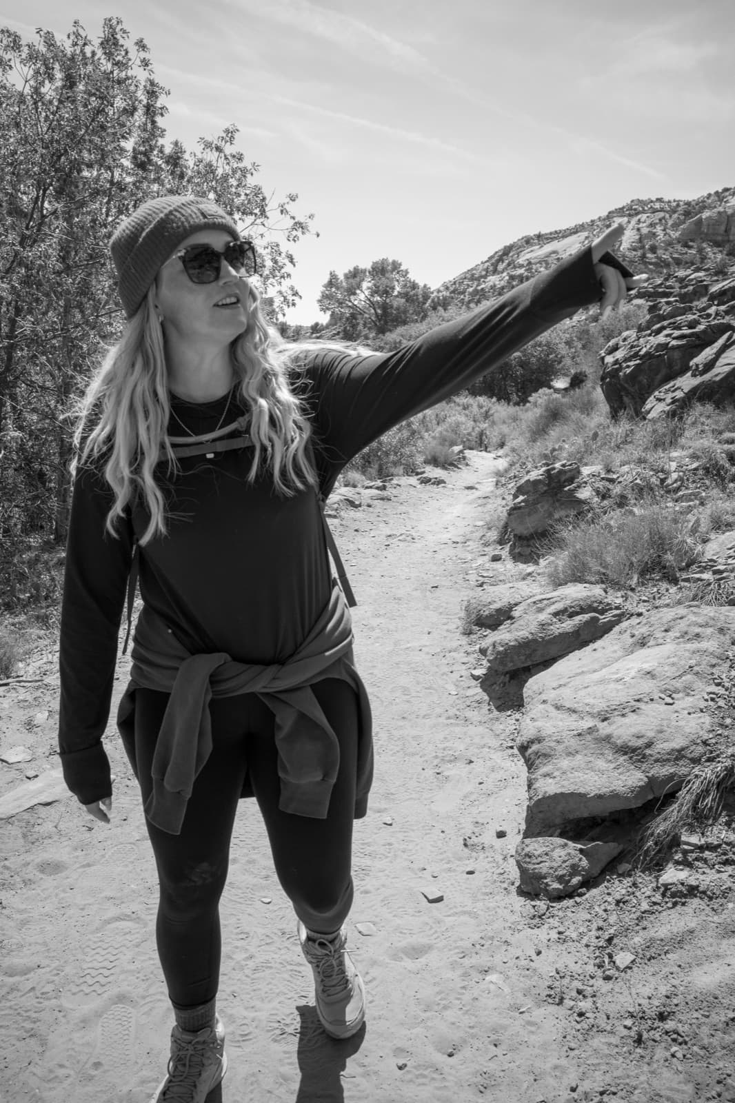 A woman points toward a rock formation while standing on a bright desert trail.