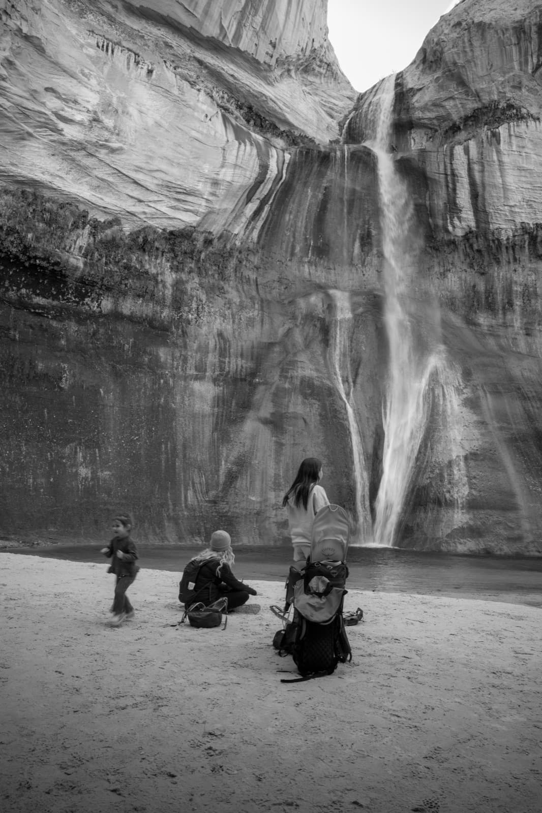 A family group stands at the base of a tall waterfall beneath towering cliffs.
