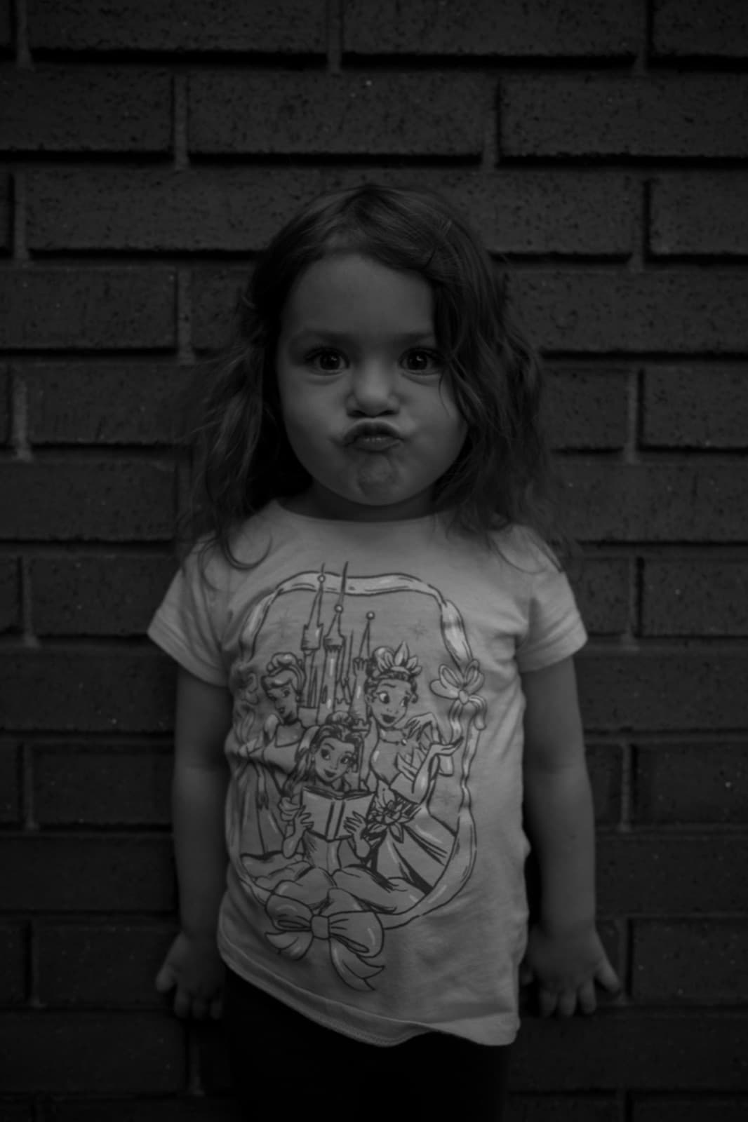 A young child makes a puckered face while standing against a dark brick wall.
