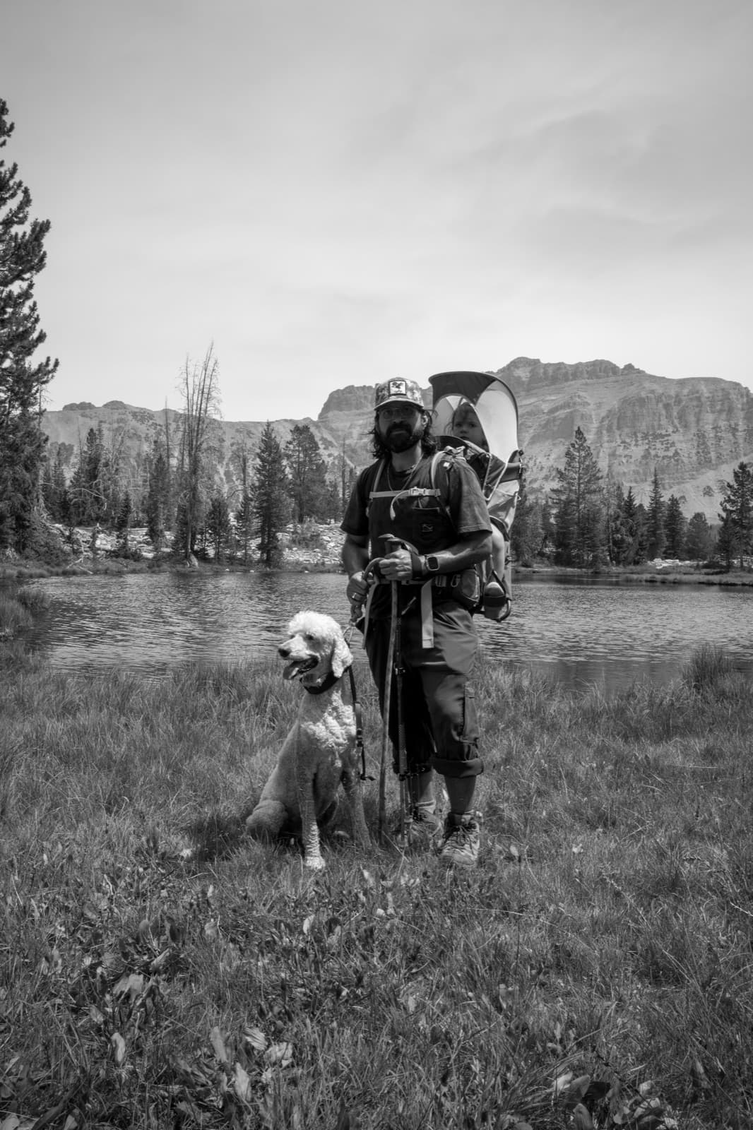 Carlos stands in a mountain meadow with a child carrier and a dog.