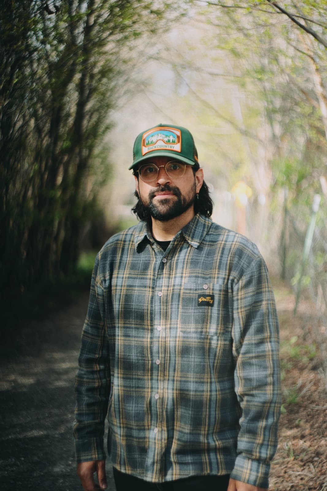 Carlos Montoya standing on a wooded trail in a green cap and plaid shirt.