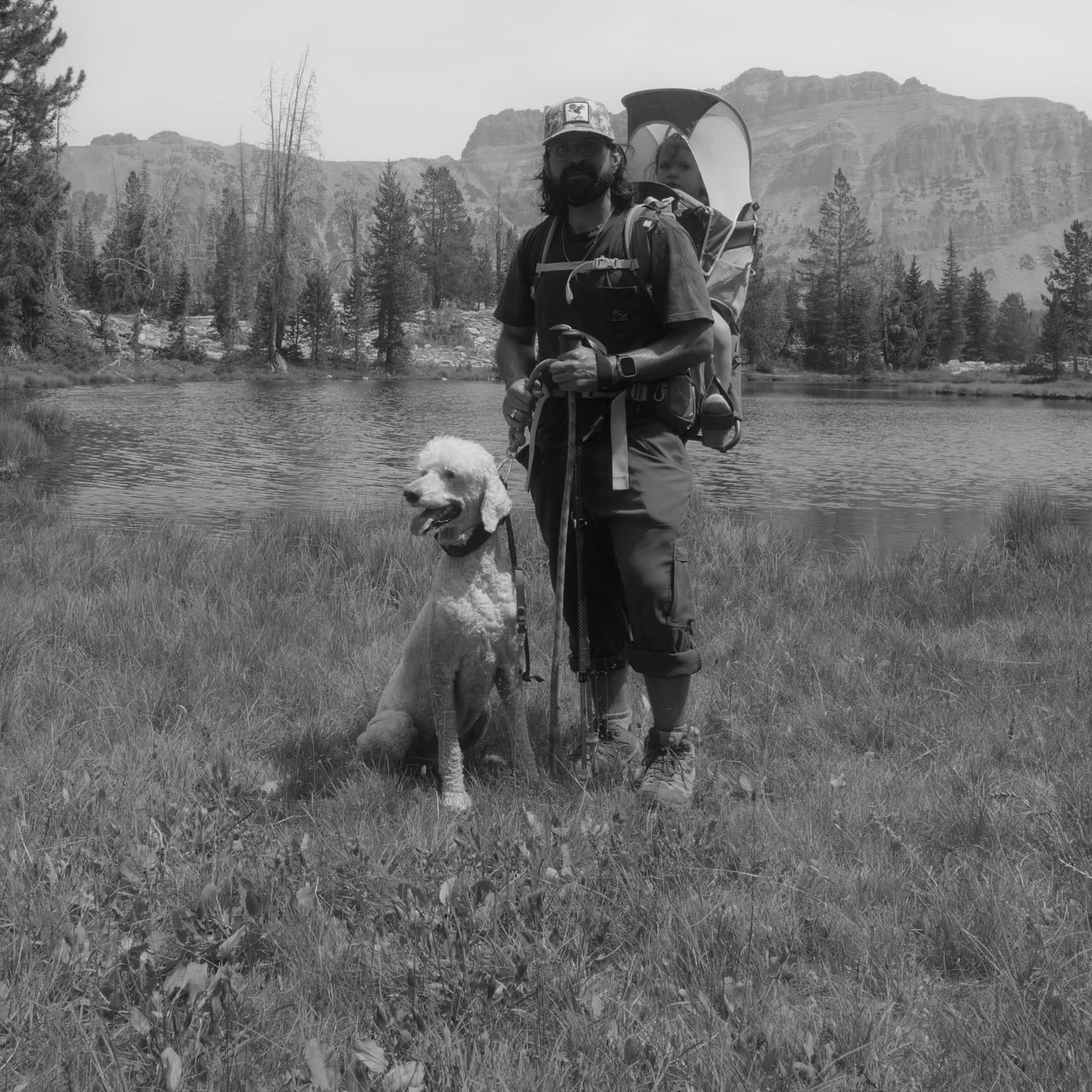 Carlos stands beside a mountain lake with a child carrier and a dog