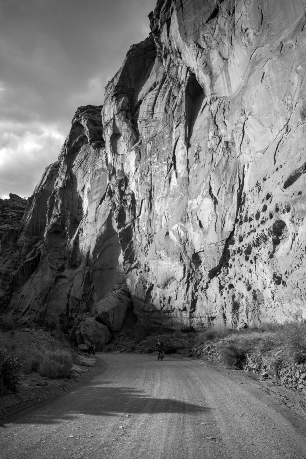 A narrow road curves beneath a tall sunlit canyon wall in a black-and-white landscape.